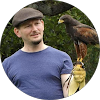 A person in a flat cap holds a brown hawk wearing a jess, standing outdoors with greenery in the background.