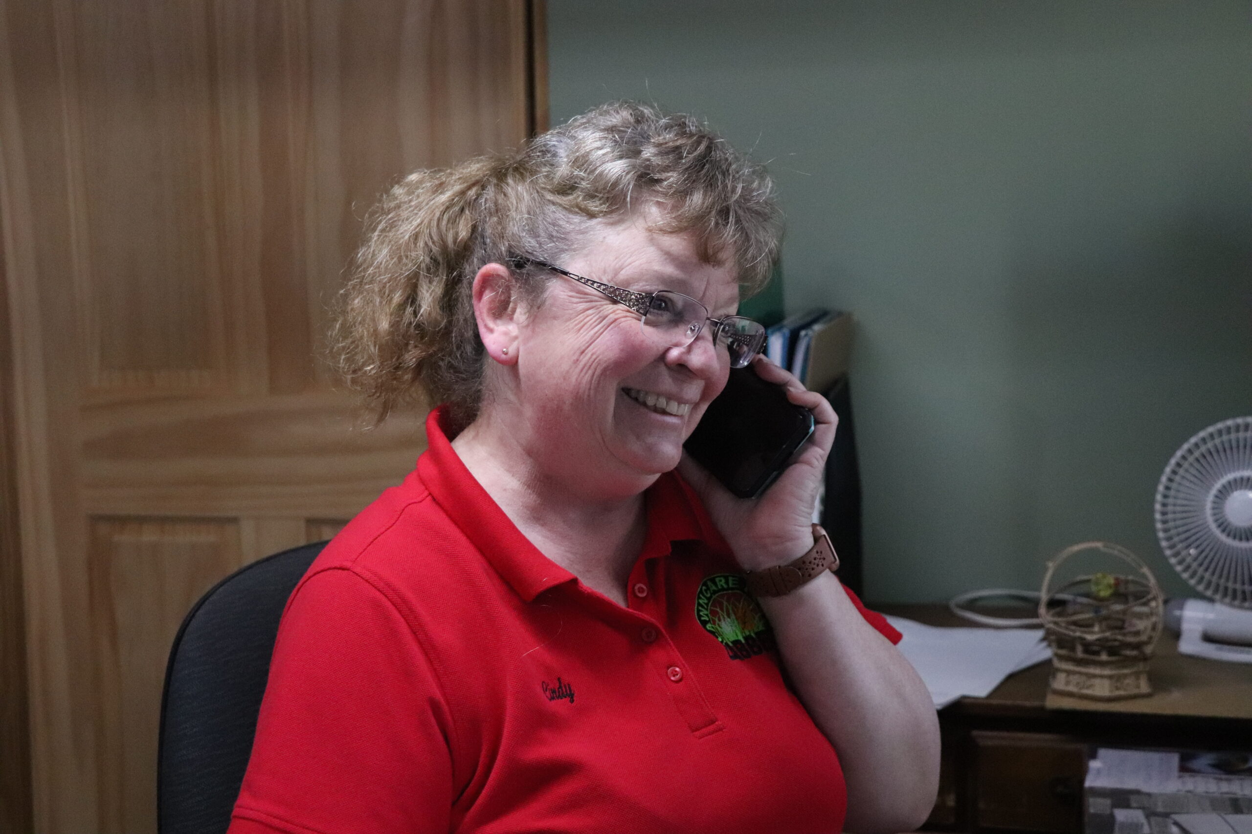 A woman wearing a red polo shirt sits at a desk and smiles while talking on a smartphone.