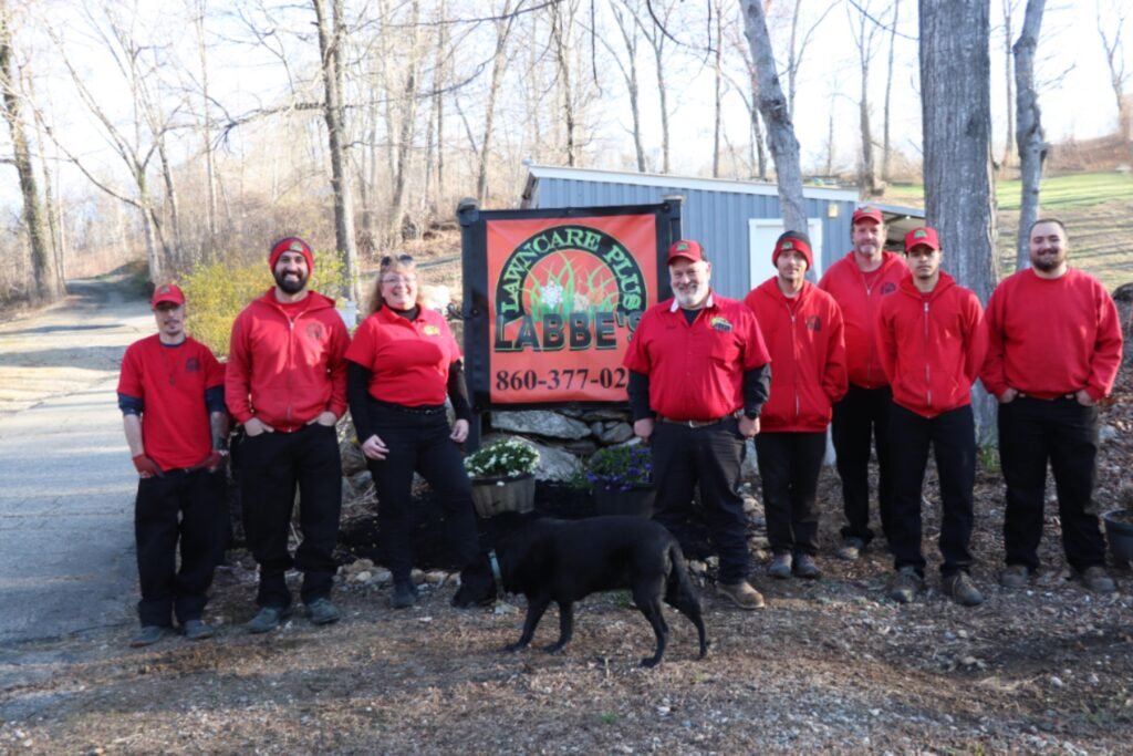 Nine people wearing red shirts and hats stand in front of a sign that reads "Mancare Hill Abbey." A black dog stands in the foreground. The background features leafless trees.