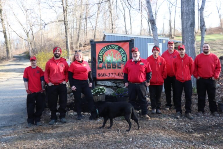 Nine people wearing red shirts and hats stand in front of a sign that reads "Mancare Hill Abbey." A black dog stands in the foreground. The background features leafless trees.