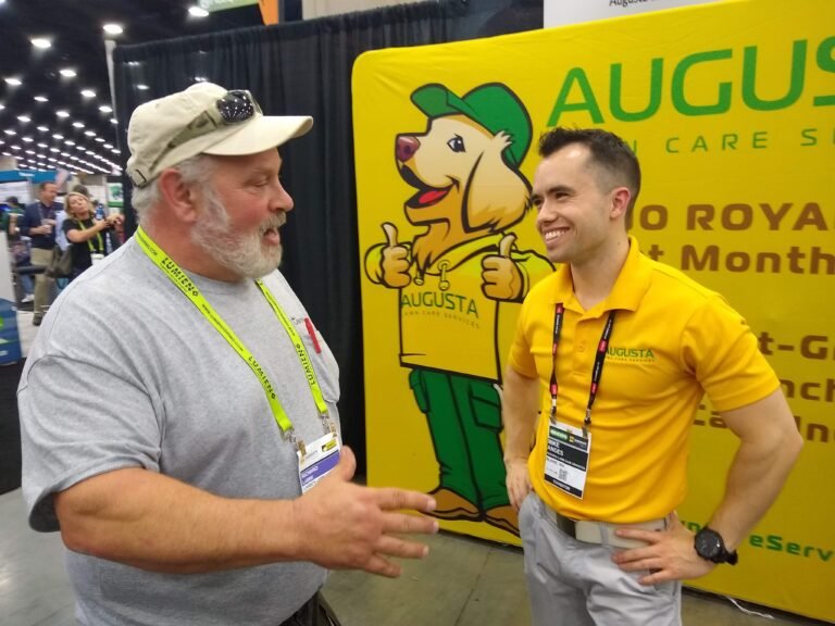 Two men converse at a trade show; one wears a gray shirt and cap, the other wears a yellow Augusta Lawn Care Service shirt, standing by a branded backdrop.