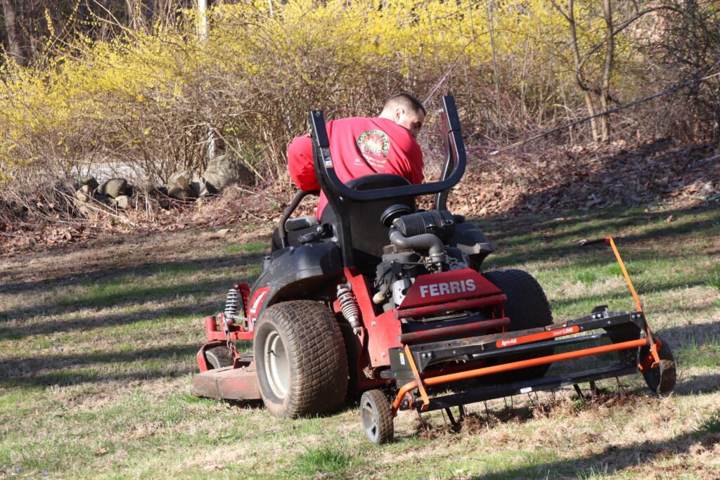 A person operates a Ferris riding lawn mower with an attached aerator, helping to dethatch the lawn in Northeast Connecticut; trees and yellow-flowering bushes provide a scenic background.