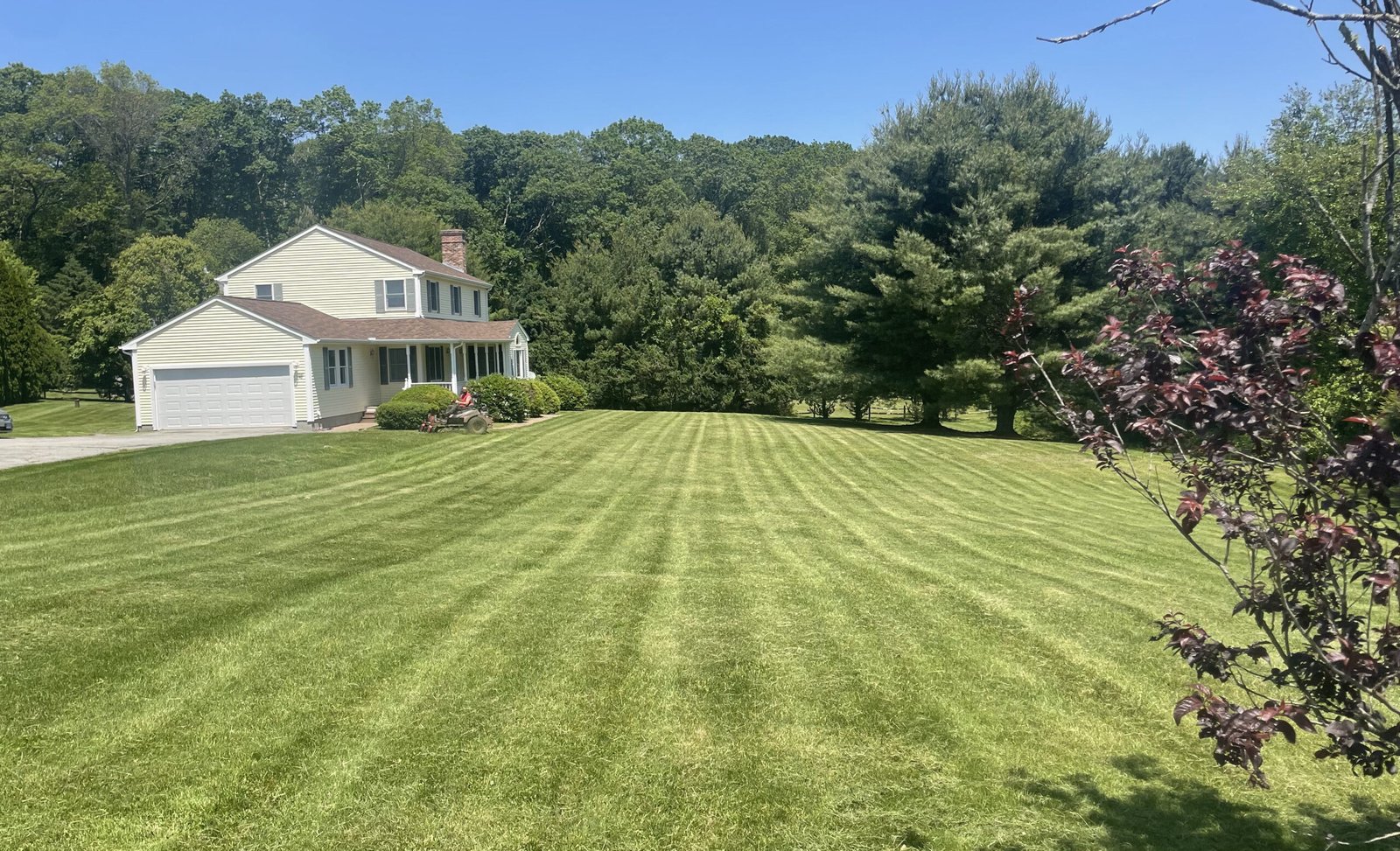 A two-story house with a large, freshly mowed lawn and surrounding trees under a clear blue sky.